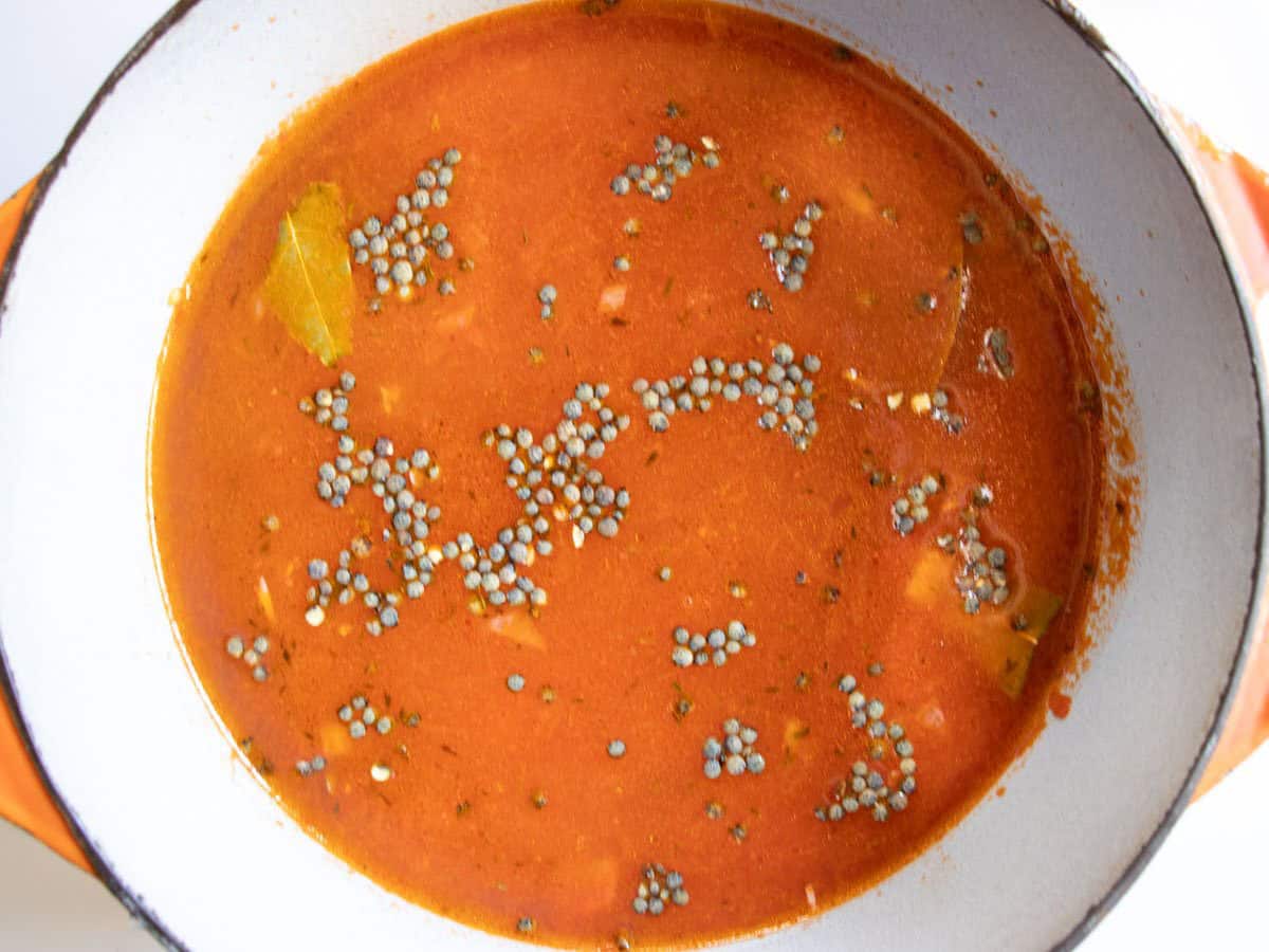 Uncooked lentils showing on surface of tomato coloured soup in overhead shot of saucepan.