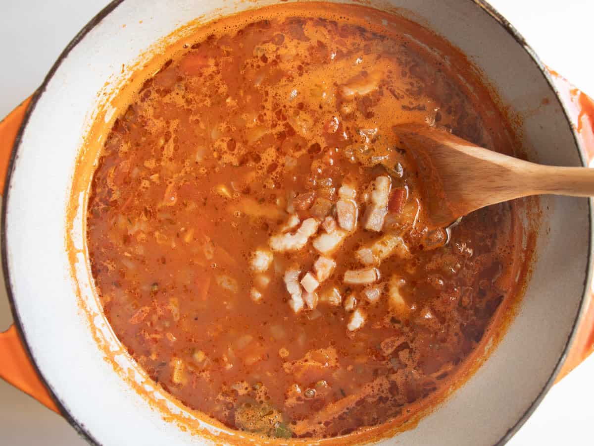 Bacon pieces have just been added to French lentil soup in enamelled cast iron saucepan and are being stirred in with wooden spoon.