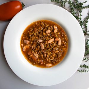 French green lentil soup in white bowl next to sprig of fresh thyme and red tomato.
