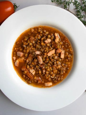 French green lentil soup in white bowl next to sprig of fresh thyme and red tomato.