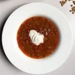 Hungarian lentil soup topped in centre with a dollop of soured cream in a white bowl next to a few scattered green lentils.