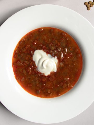 Hungarian lentil soup topped in centre with a dollop of soured cream in a white bowl next to a few scattered green lentils.