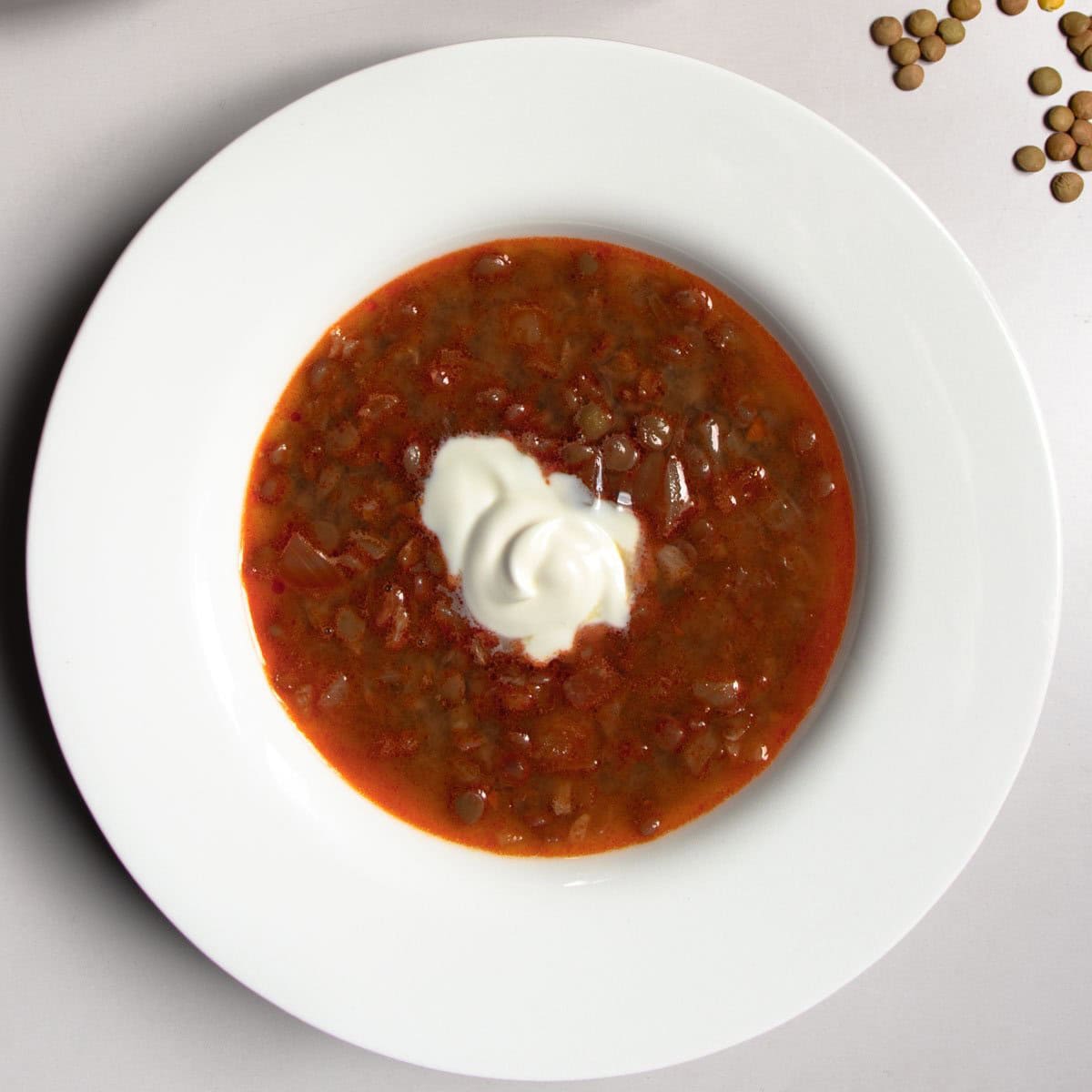 Hungarian lentil soup topped in centre with a dollop of soured cream in a white bowl next to a few scattered green lentils.