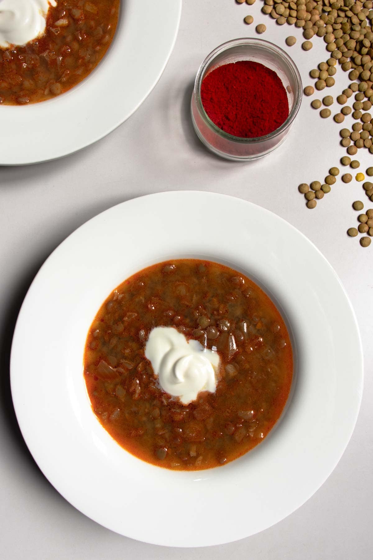 Hungarian lentil soup in white bowls next to paprika in a small glass bowl and scattered green lentils.