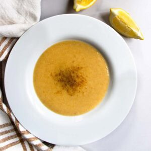 Lebanese red lentil soup in white bowl placed next to striped teacloth and slices of lemon.