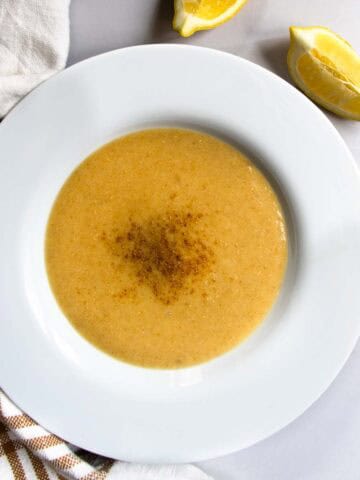 Lebanese red lentil soup in white bowl placed next to striped teacloth and slices of lemon.