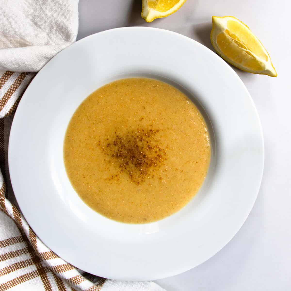 Lebanese red lentil soup in white bowl placed next to striped teacloth and slices of lemon.