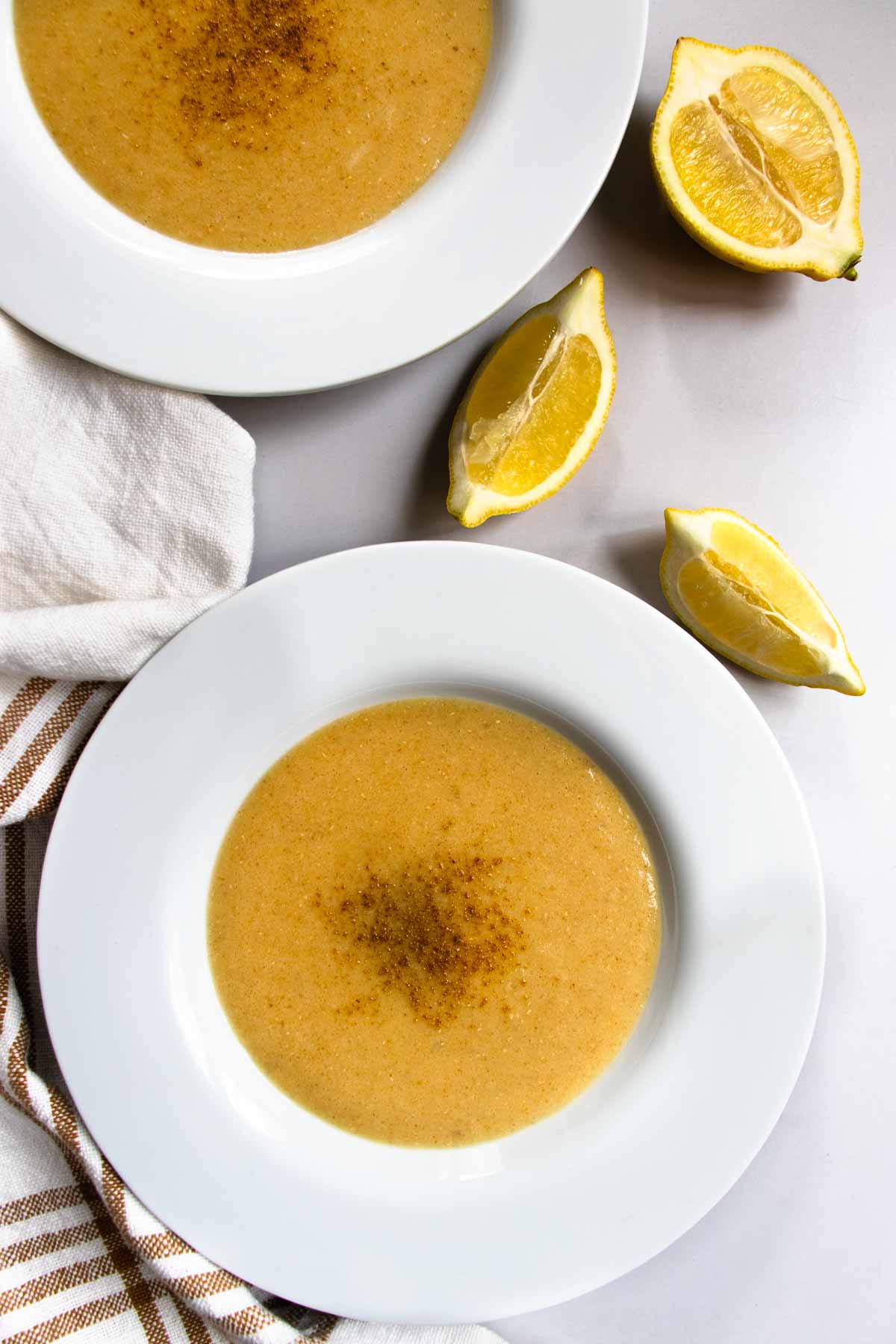 Lebanese red lentil soup in white bowls placed next to striped teacloth and lemon half and quarters.