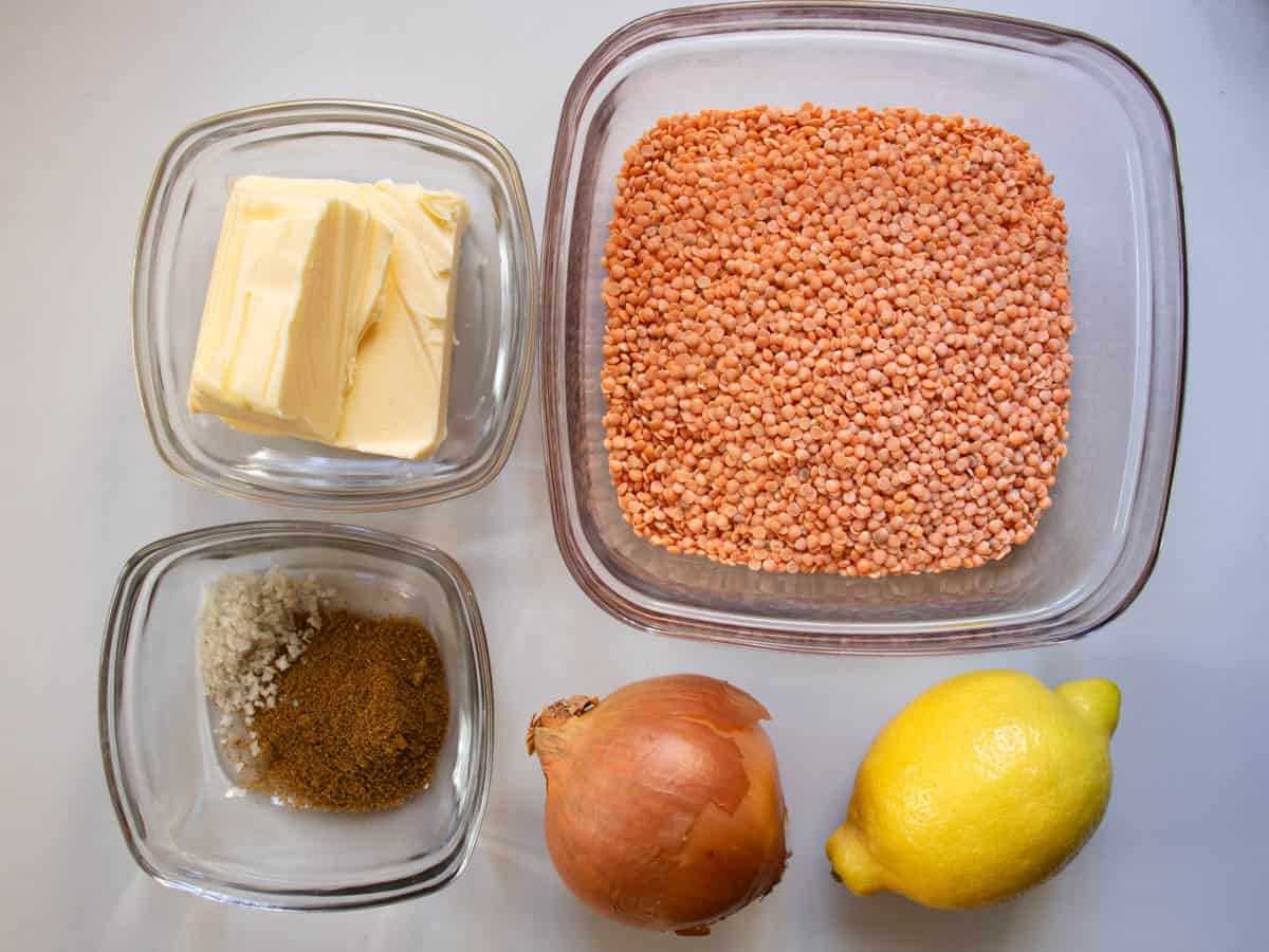 Red lentils, butter, salt and cumin all in glass bowls, an onion and lemon, all laid out on grey surface.