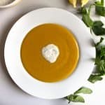 Mediterranean red lentil soup, with a dollop of yoghurt, lime and mint topping, in a white bowl next to sprig of mint and sliced lemons.