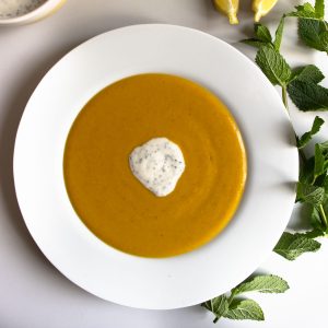 Mediterranean red lentil soup, with a dollop of yoghurt, lime and mint topping, in a white bowl next to sprig of mint and sliced lemons.