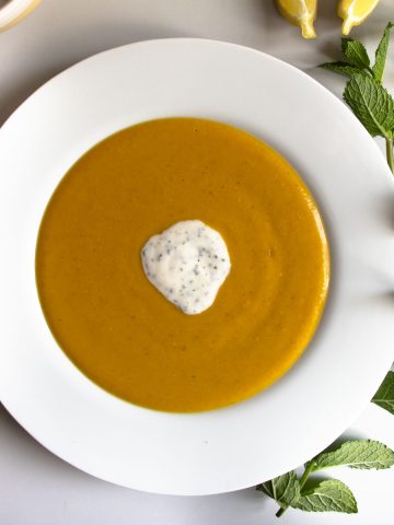 Mediterranean red lentil soup, with a dollop of yoghurt, lime and mint topping, in a white bowl next to sprig of mint and sliced lemons.