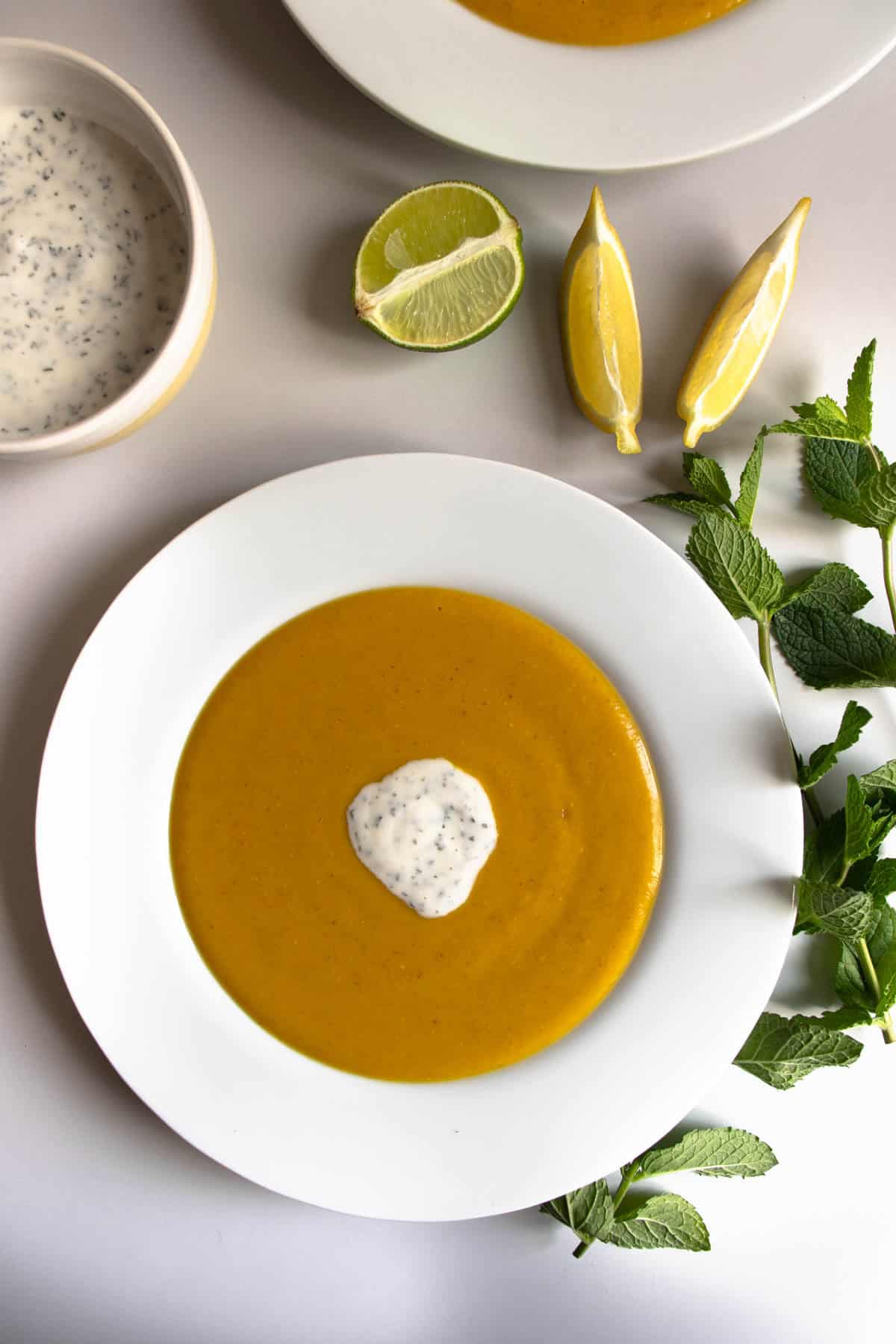 Mediterranean red lentil soup in white bowls next to sprigs of mint, sliced lemons and lime, bowl of yoghurt and mint topping.