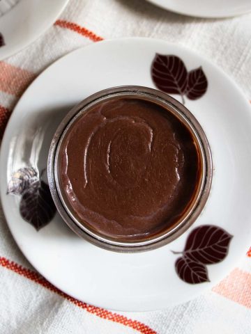 Chocolate crémeux pudding in small glass bowl on saucer with chocolate coloured leaves pattern, placed on orange red striped cloth.
