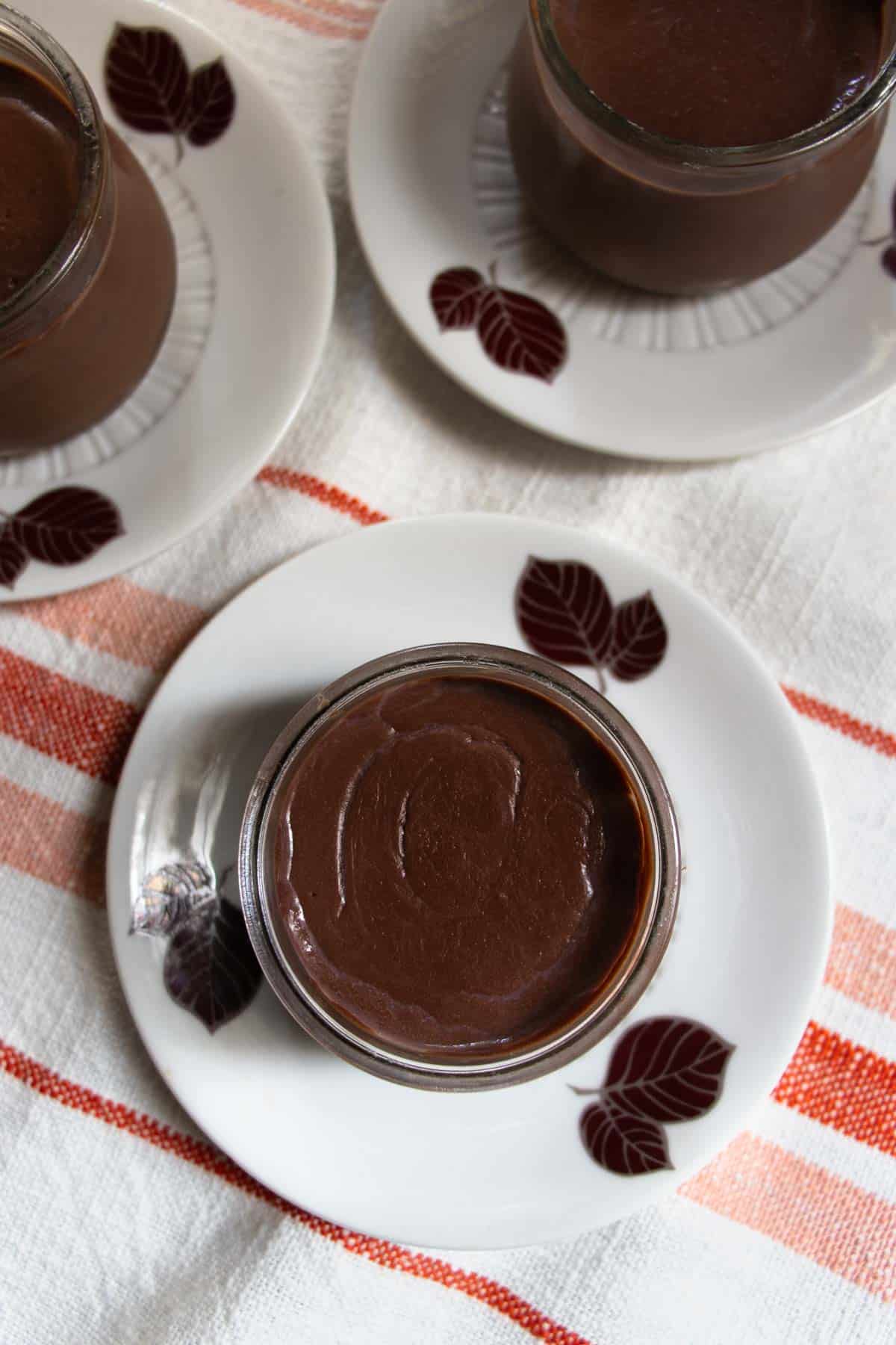 Chocolate crémeux puddings in small glass bowls placed on saucers with chocolate coloured leaves pattern, all on an orange red striped cloth.