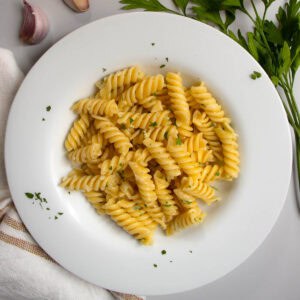 Garlic butter pasta in white dish next to cloves of garlic and sprig of flat-leaf parsley.