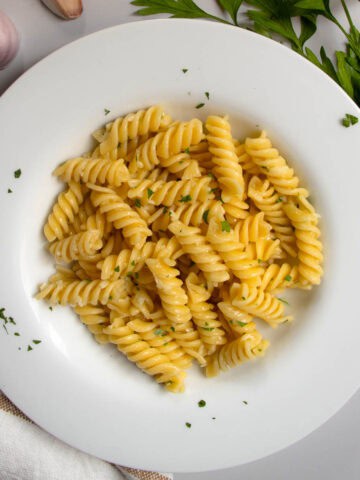 Garlic butter pasta in white dish next to cloves of garlic and sprig of flat-leaf parsley.