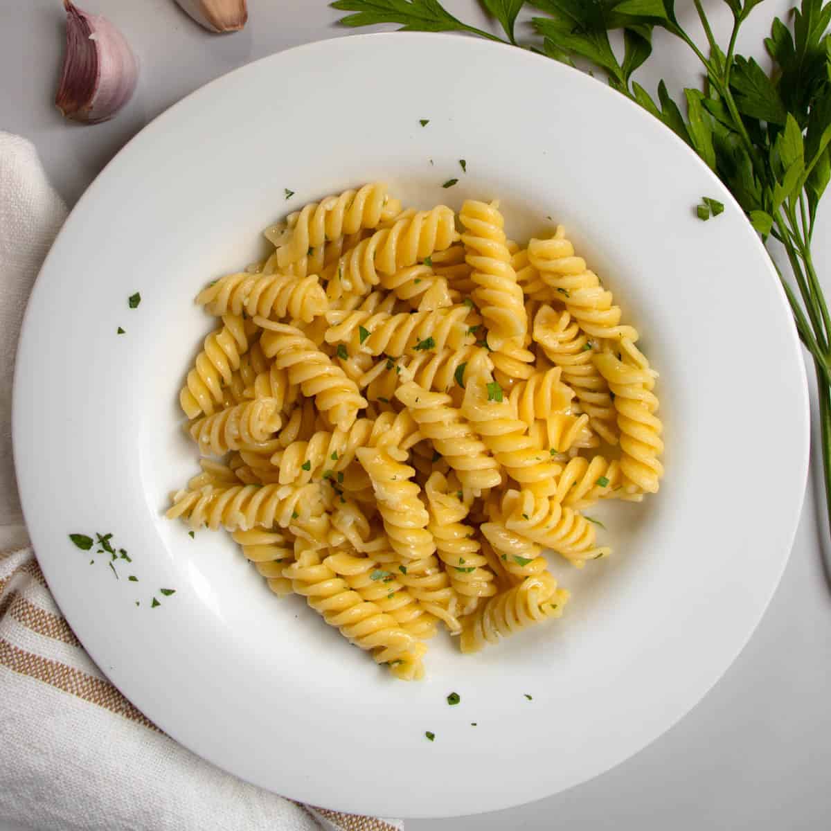 Garlic butter pasta in white dish next to cloves of garlic and sprig of flat-leaf parsley.