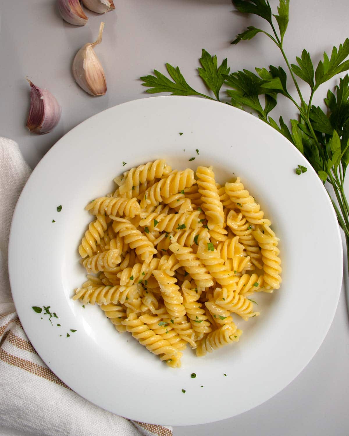 Garlic butter pasta in white dish next to cloves of garlic and sprig of flat-leaf parsley.