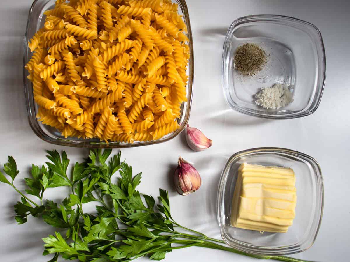 Ingredients for garlic butter pasta laid out on grey surface, pasta, garlic, butter, sprigs of parsley, salt and pepper in glass bowl.