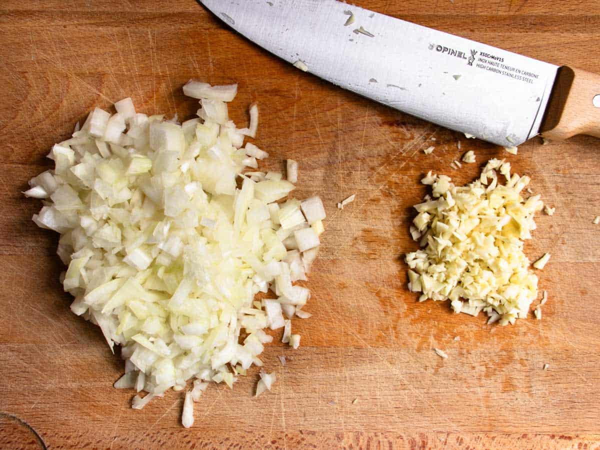 Finely chopped onion and garlic in separate piles, next to chef's knife, all on wooden chopping board.