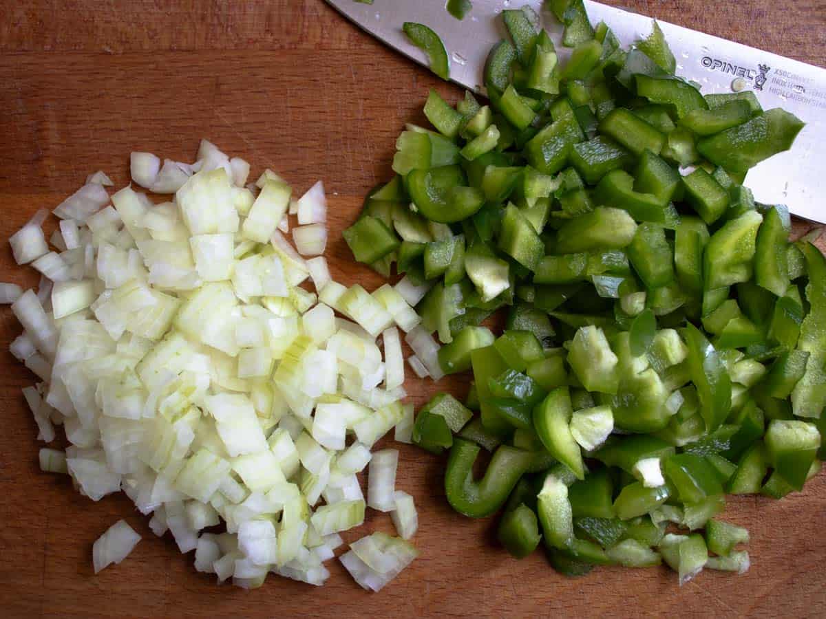 Finely chopped onions and chopped green pepper on wooden board with chef's knife.