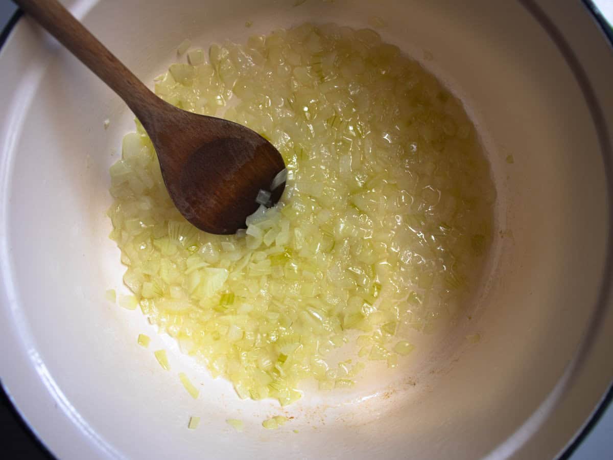 Finely chopped onions with oil in saucepan, being stirred by wooden spoon.