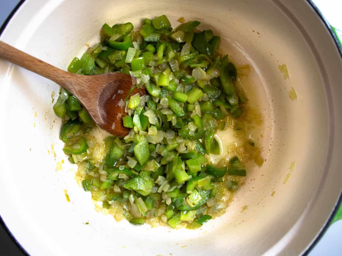 Chopped green peppers, chopped onion and cumin in saucepan being stirred by wooden spoon.