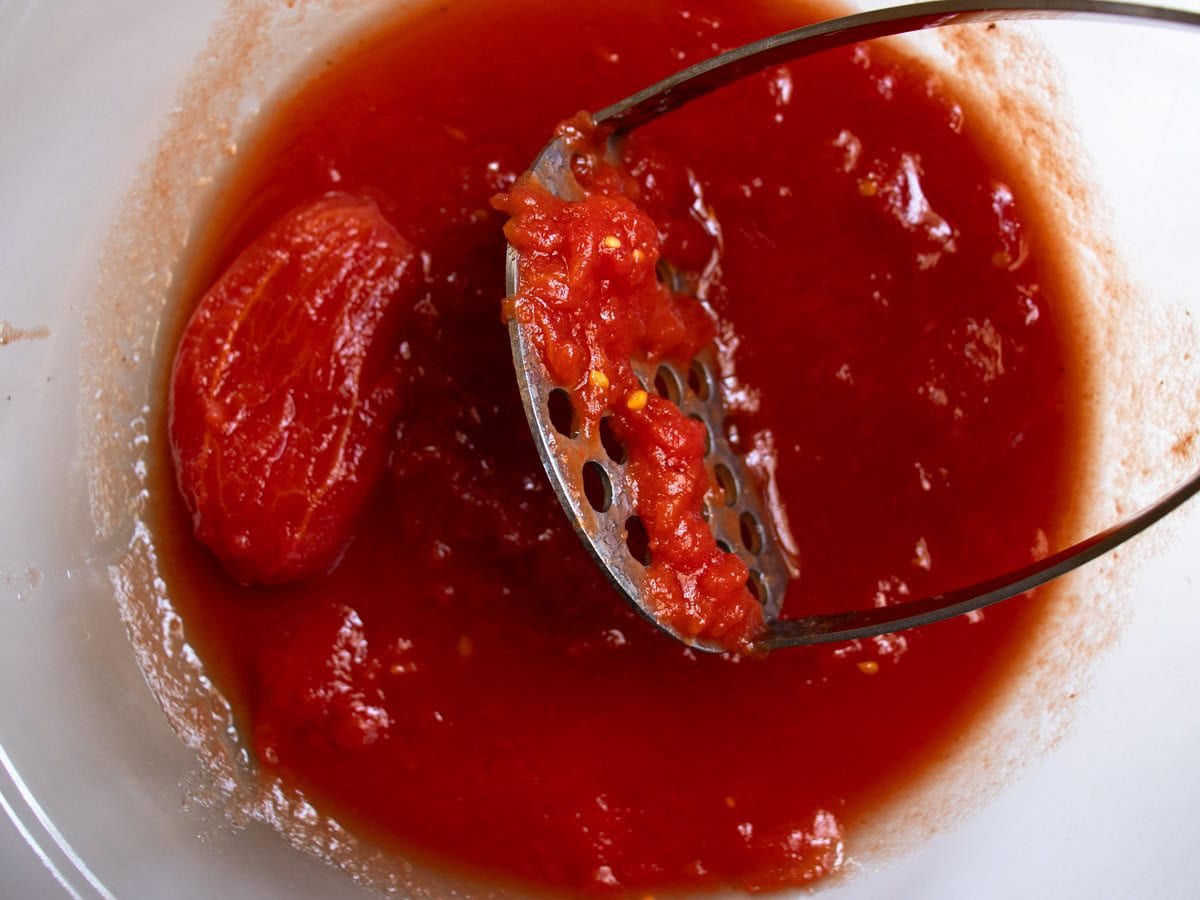 Hand held potato masher crushing whole tinned tomatoes in glass mixing bowl.