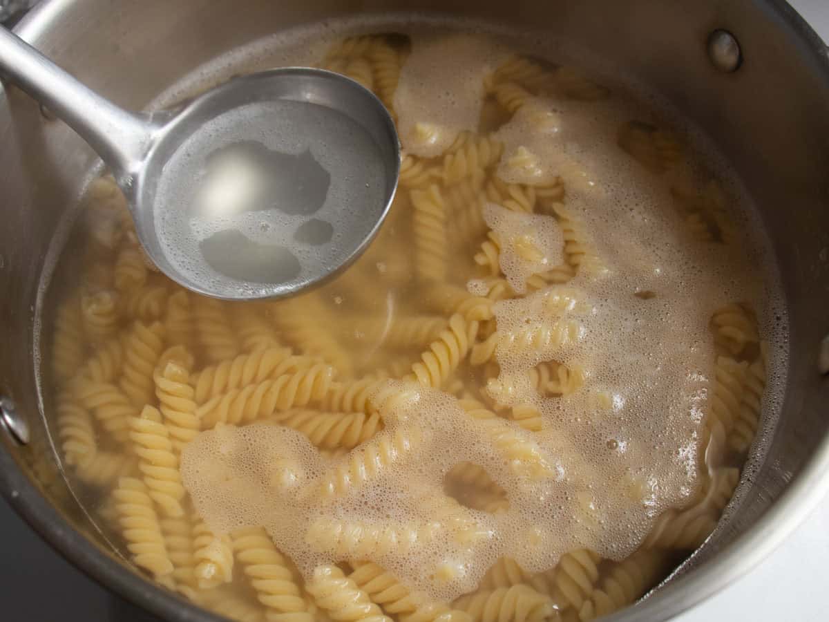 A ladle full of pasta water being held over saucepan of fusilli in hot water.