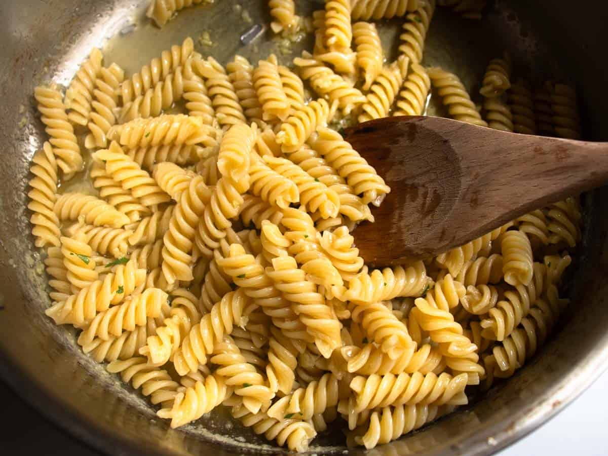 Cooked fusilli pasta being stirred with wooden spoon in butter sauce with finely chopped parsley in stainless steel saucepan.