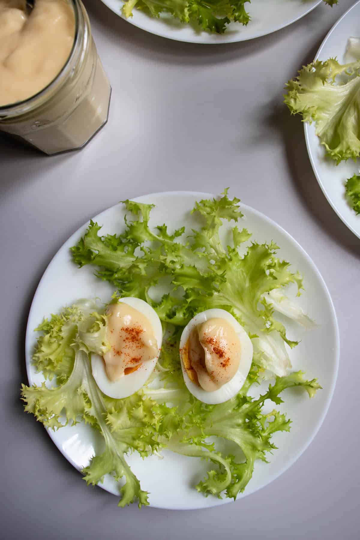 Oeufs mayonnaise sprinkled with paprika and arranged on lettuce on white plates, next to jar of mayonnaise.