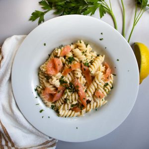 Smoked salmon creme fraiche pasta, sprinkled with chopped parsley, on white plate next to slices of lemon and sprigs of parsley.