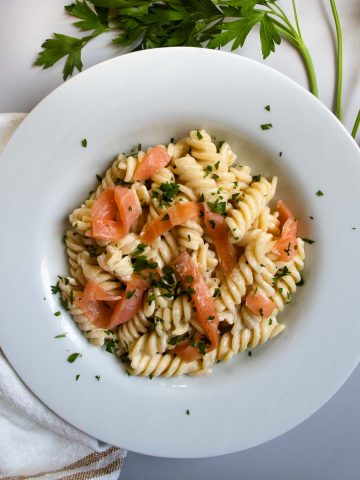 Smoked salmon creme fraiche pasta, sprinkled with chopped parsley, on white plate next to slices of lemon and sprigs of parsley.