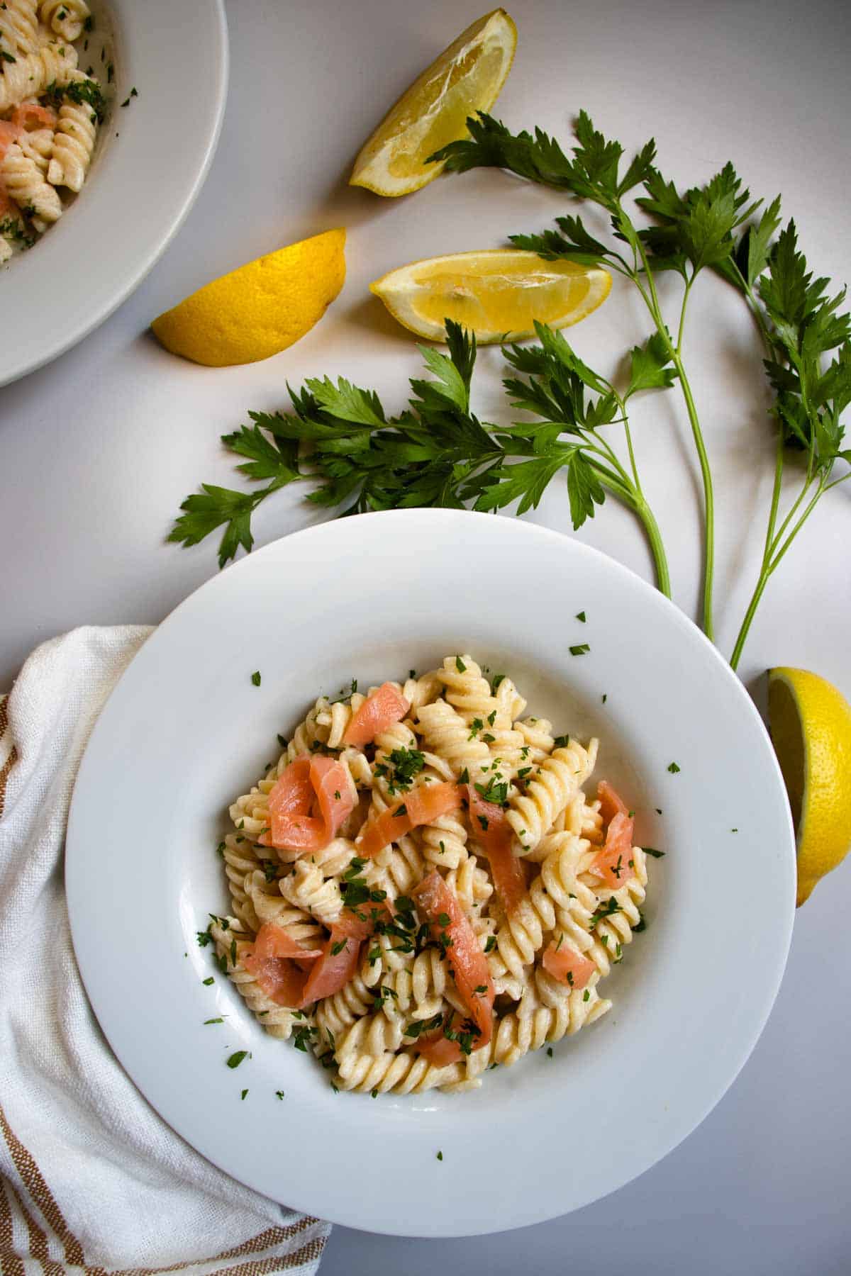 Smoked salmon creme fraiche pasta, sprinkled with chopped parsley, on white plates next to slices of lemon and sprigs of parsley.