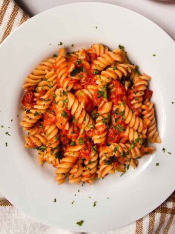 Fusilli pasta with creme fraiche and tomato sauce, sprinkled with chopped parsley in white dish, next to fresh tomatoes.