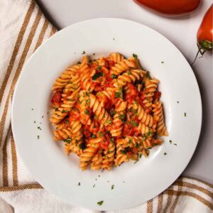 Fusilli pasta with creme fraiche and tomato sauce, sprinkled with chopped parsley in white dish, next to fresh tomatoes.