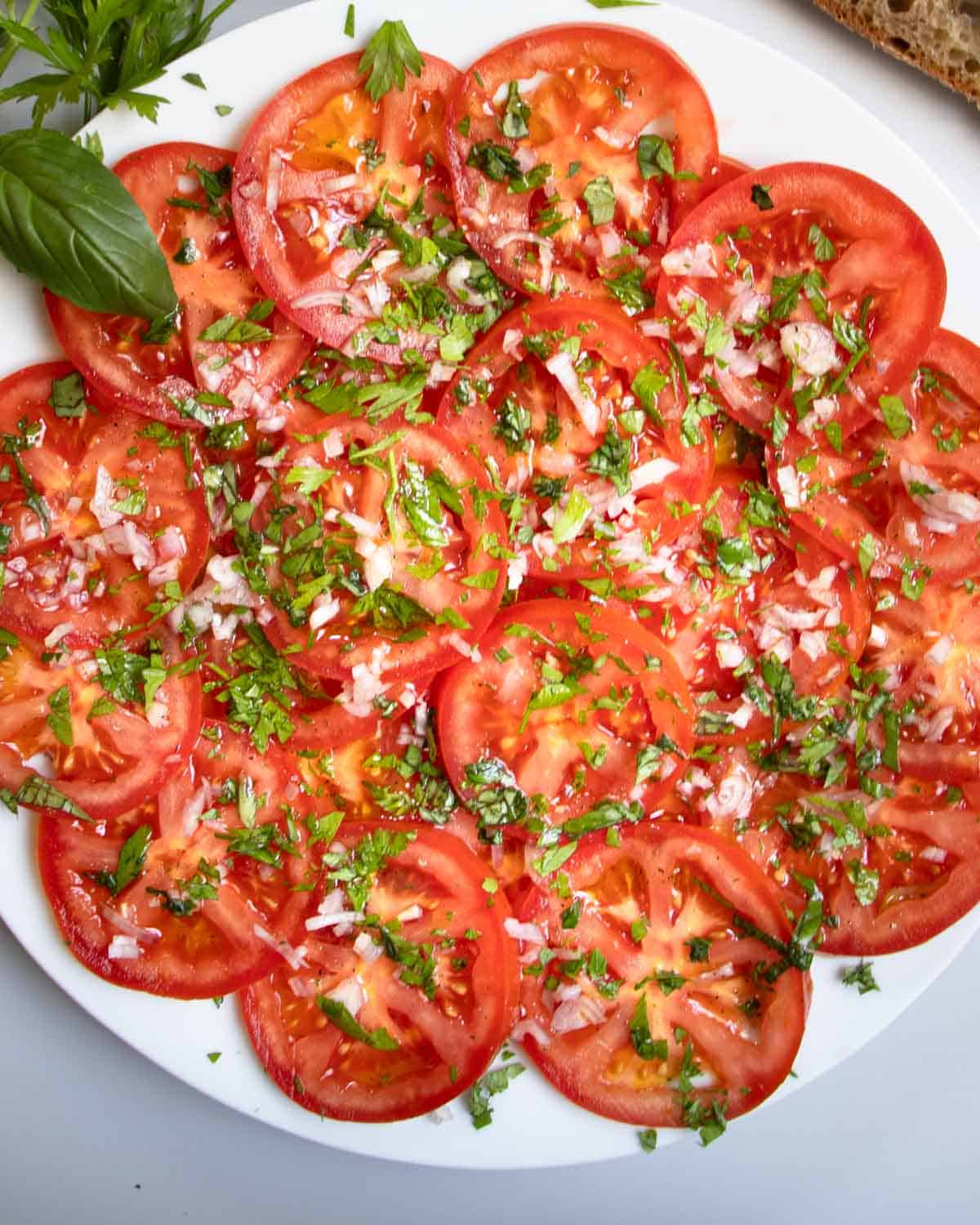 French tomato salad with circles of sliced tomatoes sprinkled with chopped parsley, basil and shallots on white plate.