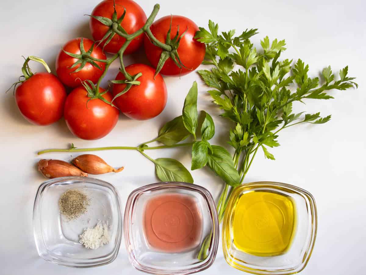 Vine tomatoes, a sprig of basil, sprigs of flat leaf parsley, a couple of shallots next to 3 glass dishes with vinaigrette dressing ingredients.