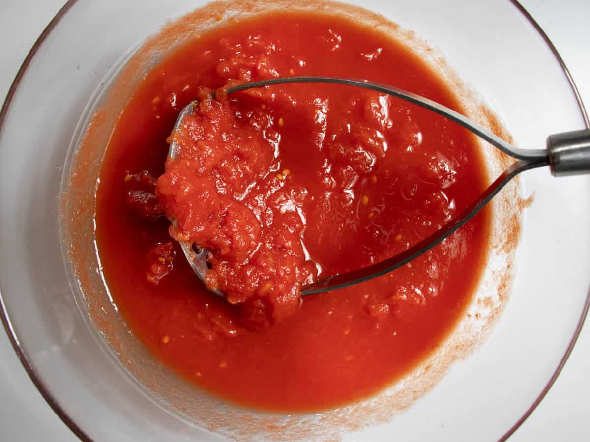 Canned tomatoes in glass bowl being crushed by potato masher.