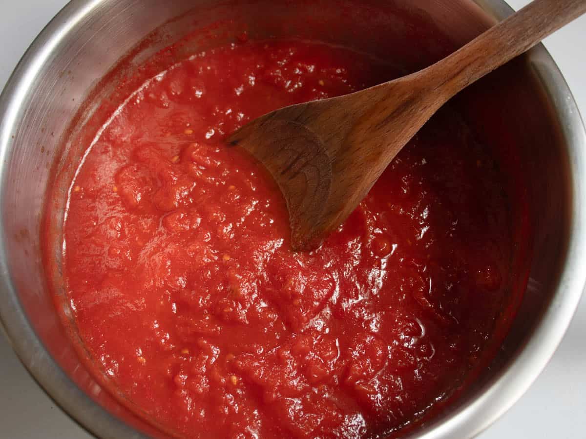 Tomato sauce in stainless steel saucepan being stirred with wooden spoon.