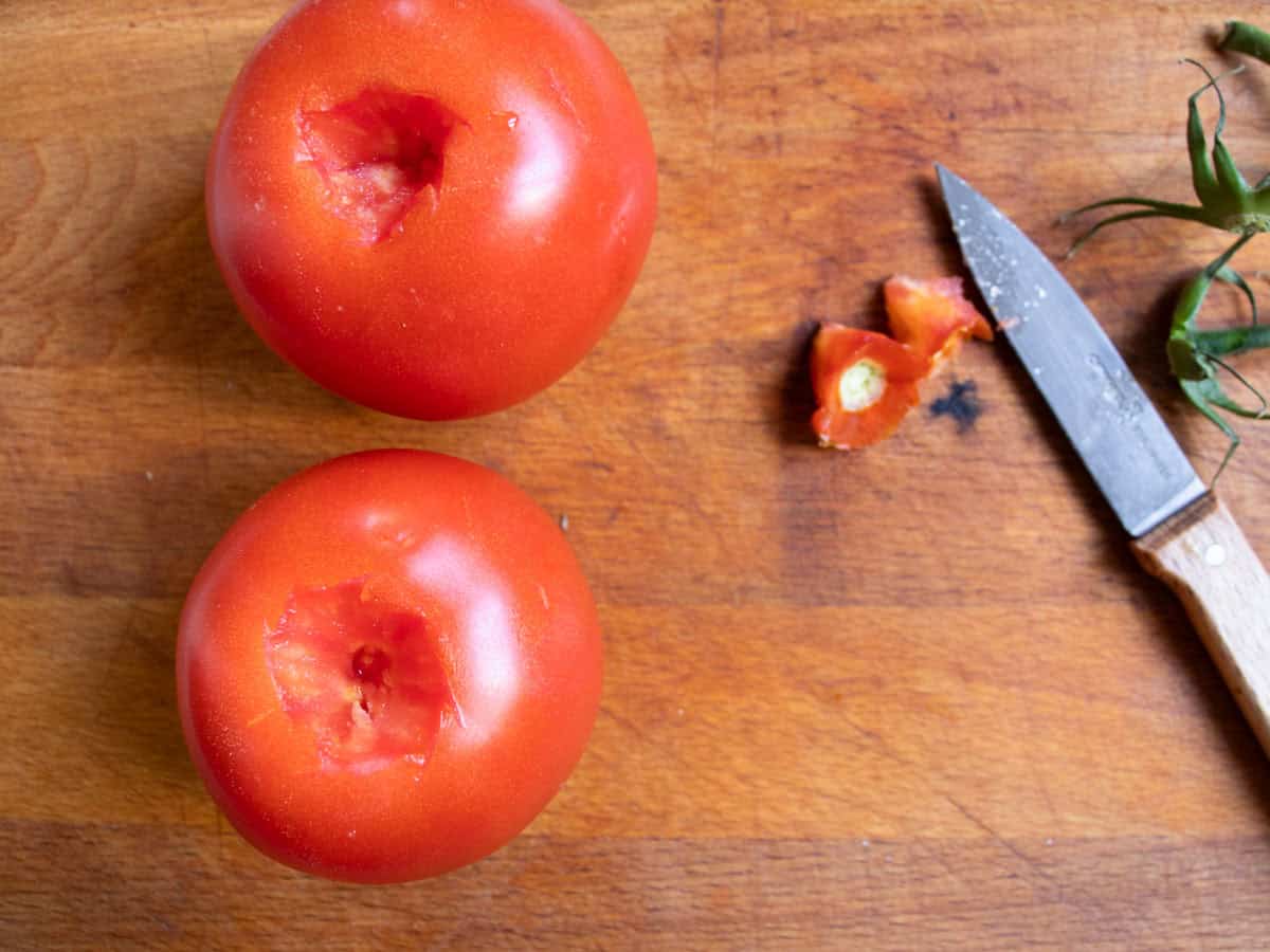 Two tomatoes on wooden board with core top removed, removed cores next to small sharp knife.
