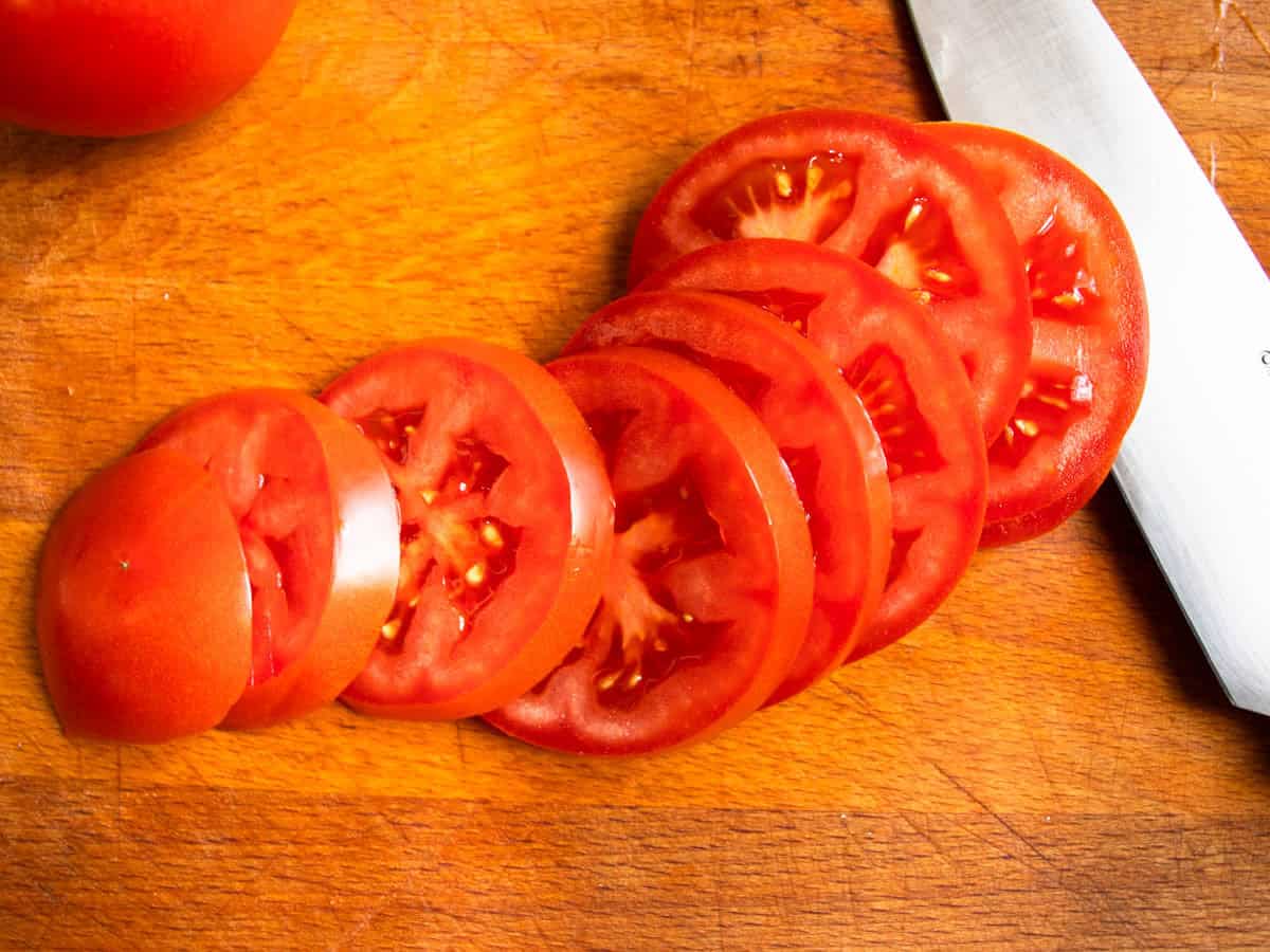 A tomato sliced in rounds on wooden board, next to chef's knife.
