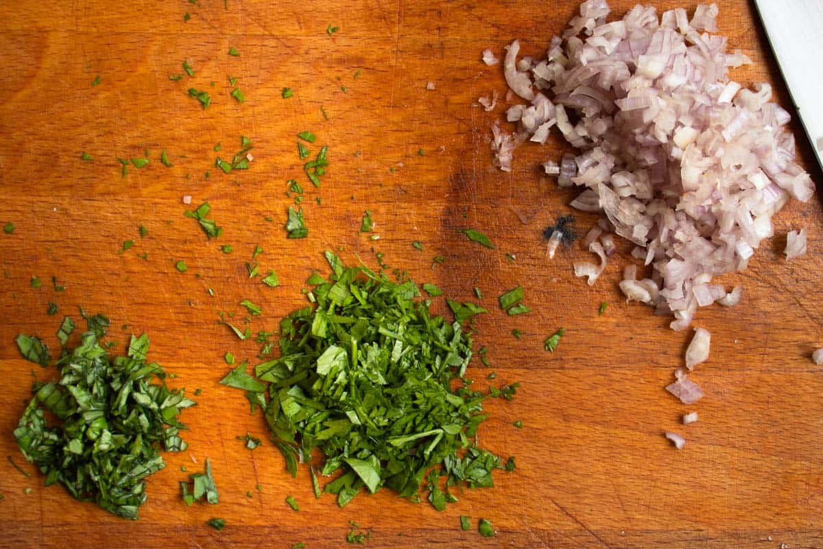 Finely chopped parsley, basil and shallots in small mounds on wooden board.