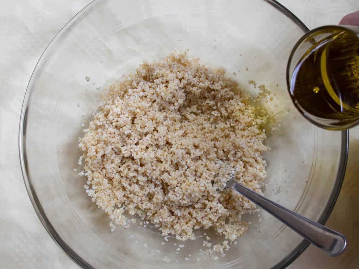 Olive oil being poured from glass onto lemon-soaked bulgur wheat in glass mixing bowl.