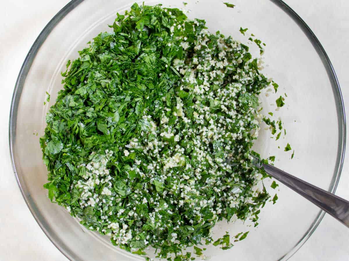 Chopped parsley and mint being mixed together in glass mixing bowl with bulgur wheat by stainless steel spoon.