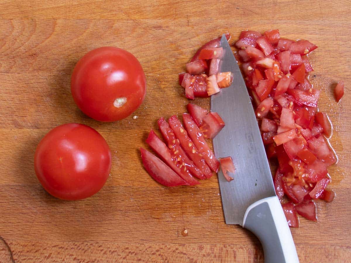 Two tomatoes on a wooden board, a third tomato being cut into small dice with chef's knife.