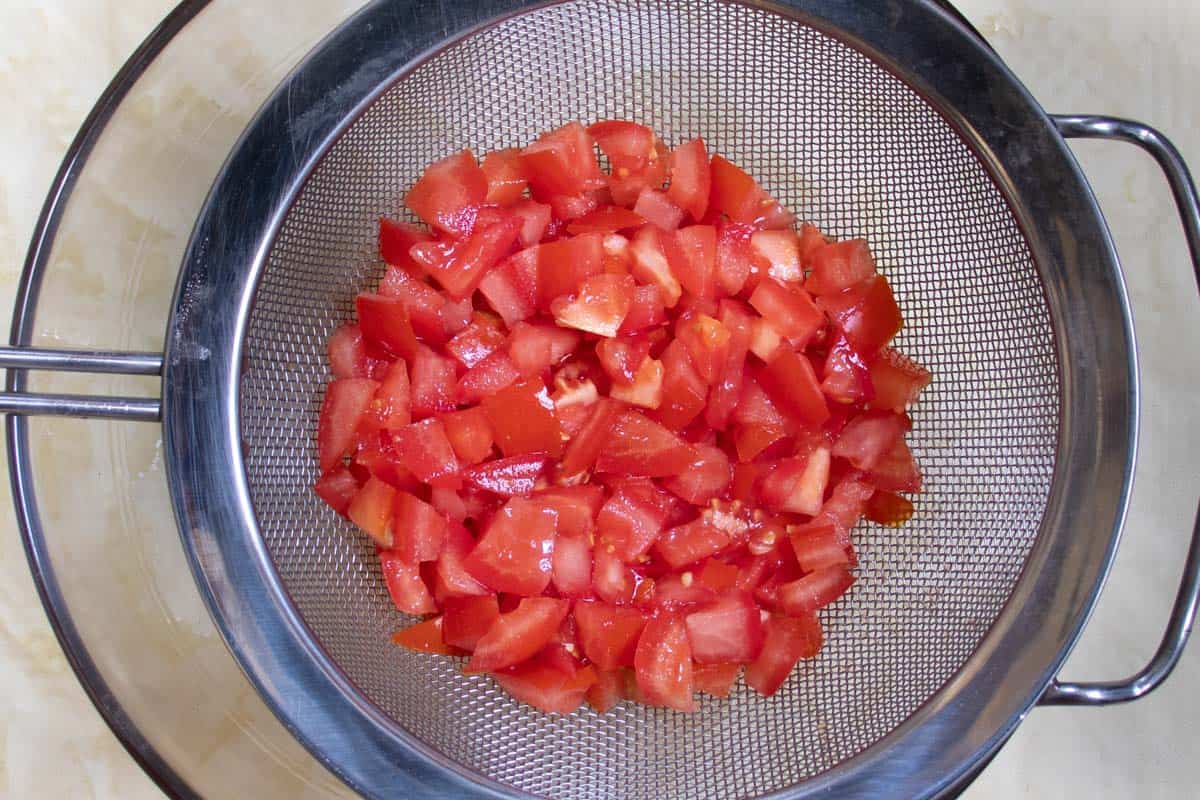 Diced tomatoes draining in a metal sieve suspended over a glass bowl.