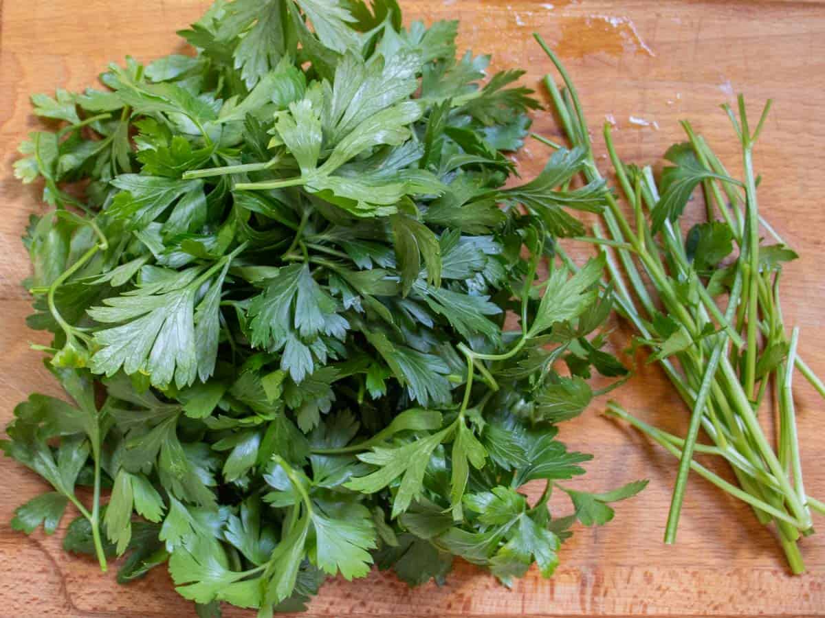 De-stemmed parsley leaves next to pile of stems on wooden board.