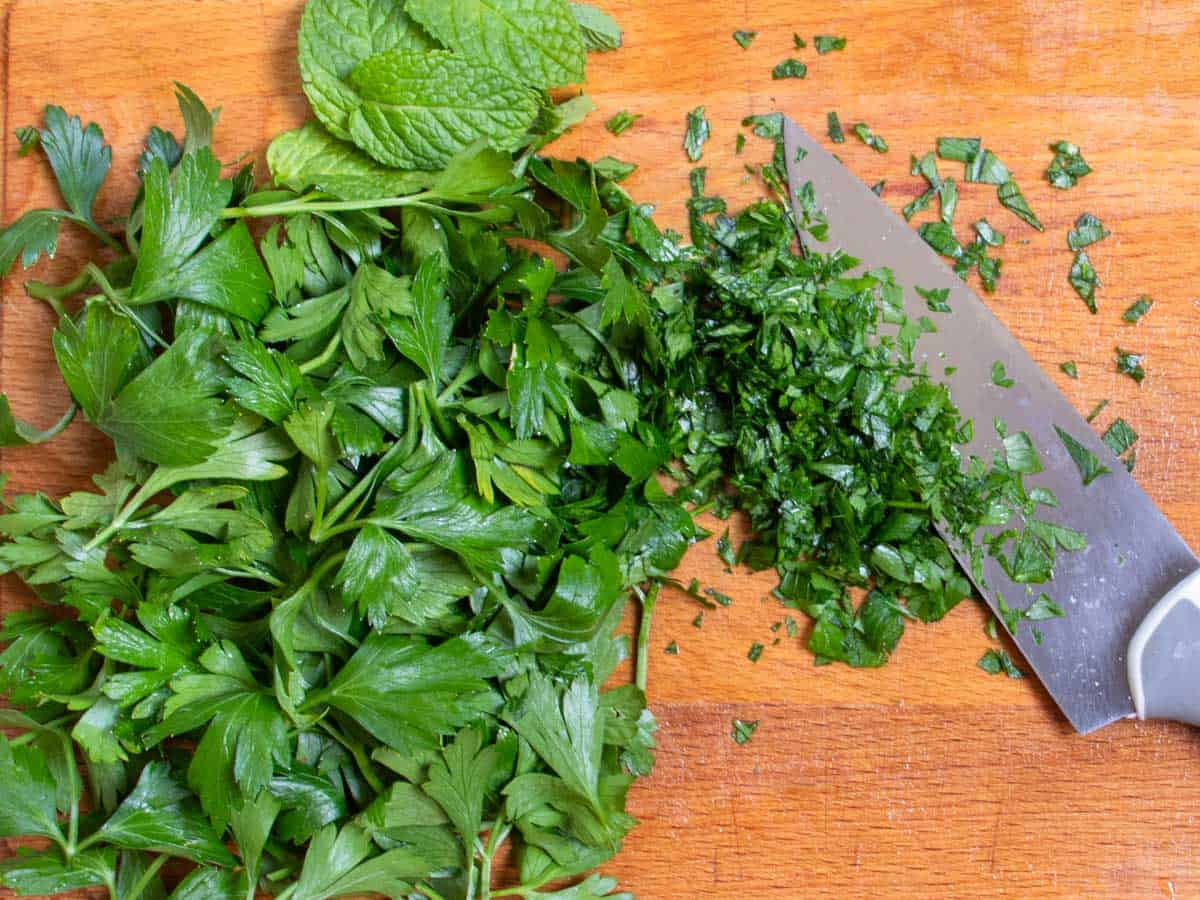 Parsley and mint leaves being chopped with chef's knife on wooden chopping board.